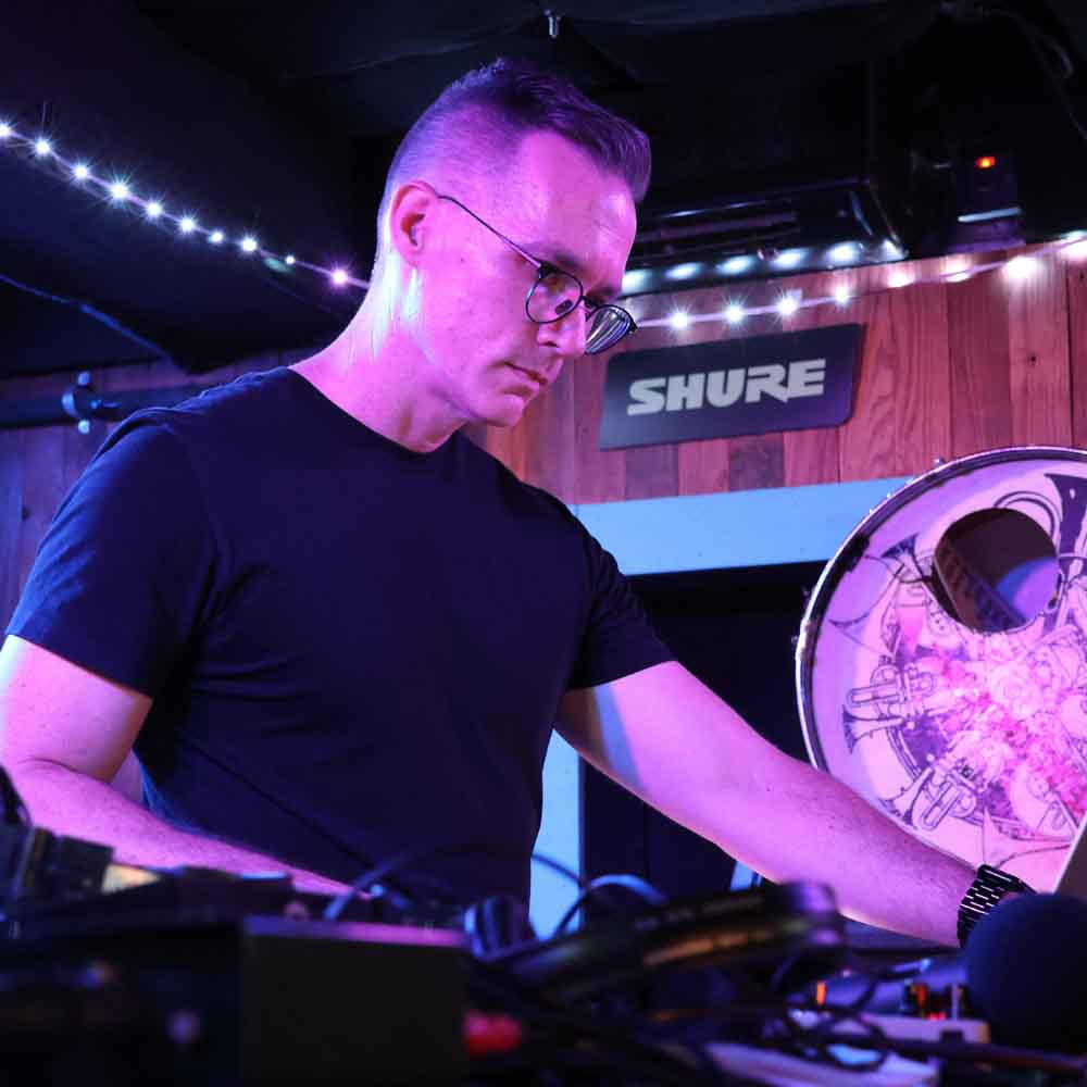 Photograph of Timothy Fairless performing live music. Shot from the side, it shows the performer and a laptop computer adorned with stickers from a cafe called Mum's Not Home. In the background is a decorative kick drum, not used for performance.